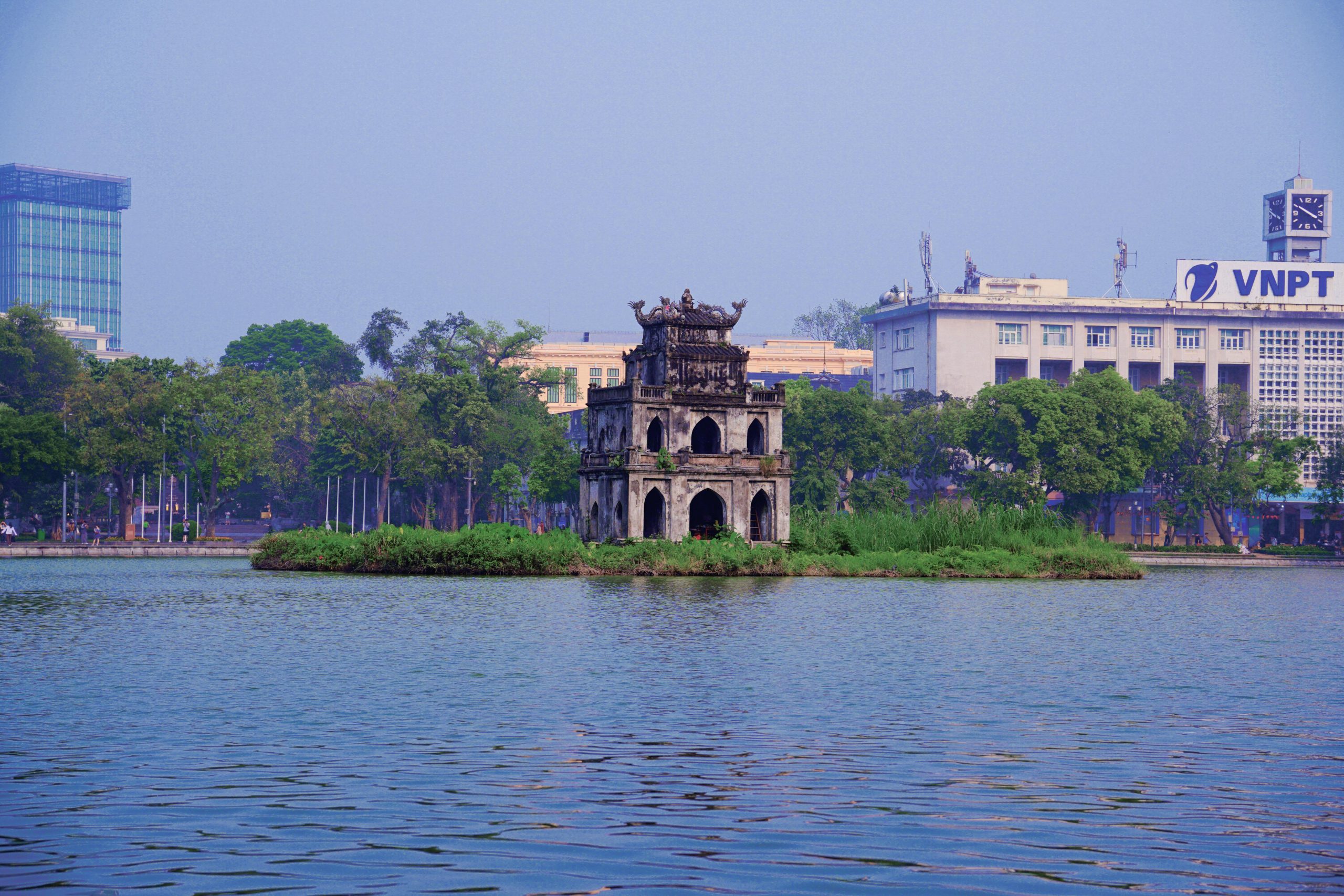 Scenic view of Turtle Tower in Hoan Kiem Lake, Hanoi, framed by greenery and urban backdrop.
